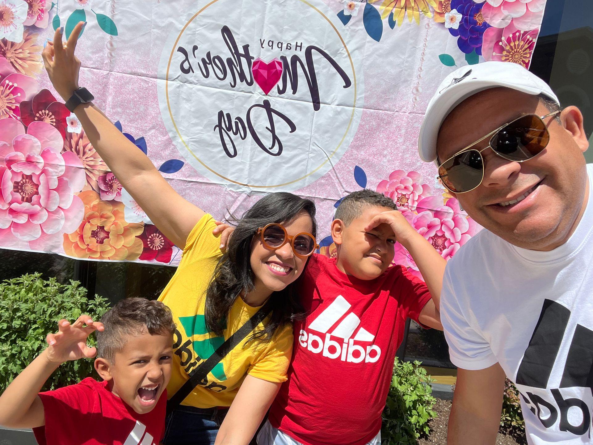 Family posing in front of a floral Mother's Day banner outside on a sunny day.