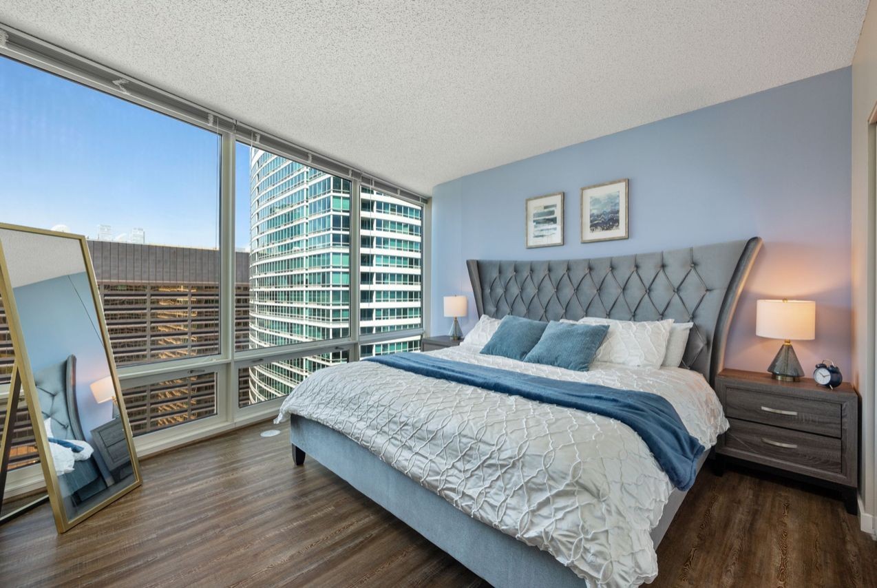 Modern bedroom with large windows, gray headboard bed, and wooden floor.